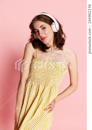 Portrait of young beautiful girl in cute dress listening to music in headphones and smiling against pink studio background 104902336