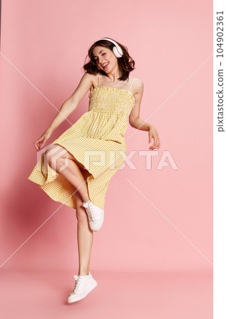 Happy, positive young pretty girl in yellow dress listening to music in headphones and dancing against pink studio background Happy, positive young pretty girl in yellow dress listening to music in headphones and dancing against pink studio background 104902361