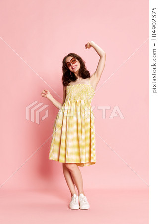 Full-length portrait of beautiful young woman in pretty yellow dress and sunglasses cheerfully dancing against pink studio background Full-length portrait of beautiful young woman in pretty yellow dress and sunglasses cheerfully dancing against pink studio background 104902375