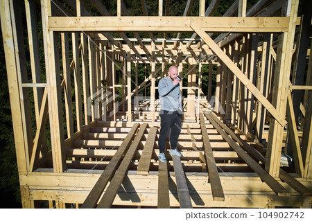 Client is having wooden frame house built near the forest, drinking a cup of coffee. The concept behind it is modern and environmentally friendly construction. 104902742