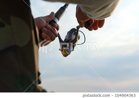 Old man fishing. Senior gray haired fisherman throws a spinning from shoreside at sunset, twisting a coil. Positive elderly male angling at lake, rotating reel rod. Retired fisher enjoying his weekend 104904363