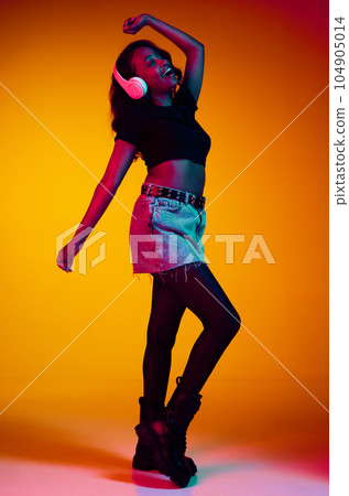 Full-length portrait of beautiful, young, african, stylish woman listening to music in headphones and dancing against yellow studio background in neon light Full-length portrait of beautiful, young, african, stylish woman listening to music in headphones and dancing against yellow studio background in neon light 104905014