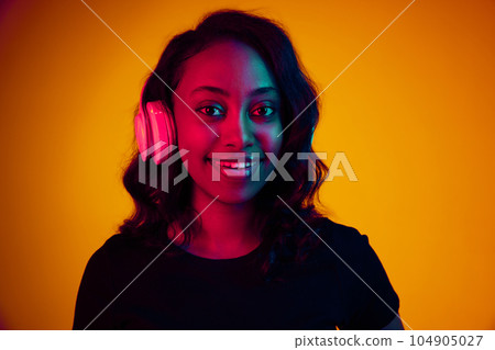 Portrait of young, beautiful, smiling african woman listening to music in headphones against yellow studio background in neon light Portrait of young, beautiful, smiling african woman listening to music in headphones against yellow studio background in neon light 104905027
