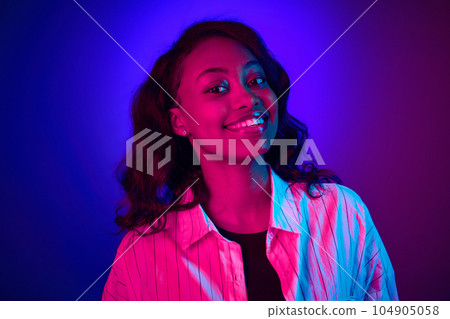 Close-up portrait of beautiful, young, african woman looking at camera, smiling against gradient blue purple studio background in neon light Close-up portrait of beautiful, young, african woman looking at camera, smiling against gradient blue purple studio background in neon light 104905058