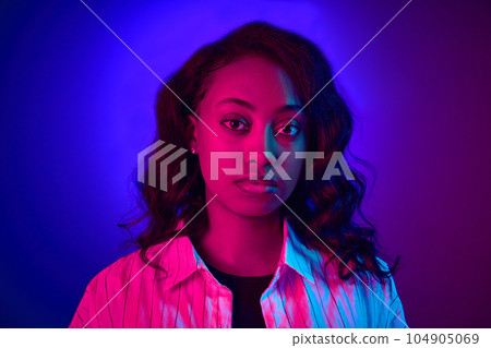Close-up portrait of beautiful, young, african woman looking at camera, posing against gradient blue purple studio background in neon light 104905069