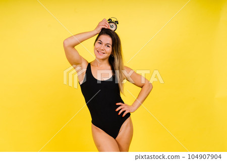 Female holding alarm clock, isolated on yellow. Young model in black swimsuit. Female holding alarm clock, isolated on yellow. Young model in black swimsuit. 104907904