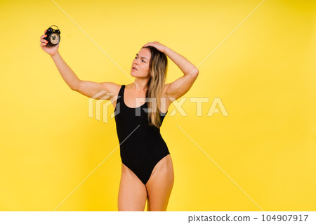 Female holding alarm clock, isolated on yellow. Young model in black swimsuit. 104907917