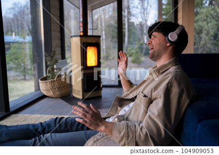 Energetic, positive guy sings loudly while listening to music with headphones, dressed in a beige shirt, sitting by the sofa in the living room against the background of the fireplace Energetic, positive guy sings loudly while listening to music with headphones, dressed in a beige shirt, sitting by the sofa in the living room against the background of the fireplace 104909053