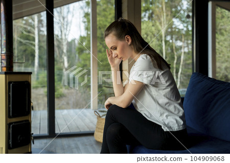 An unhappy young woman is sitting at home on a blue sofa and looking out the window, thinking about problems and troubles, suffering from depression or loneliness 104909068