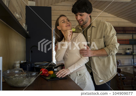 Smiling man looks at his wife, who is cooking dinner on the stove in the kitchen Smiling man looks at his wife, who is cooking dinner on the stove in the kitchen 104909077
