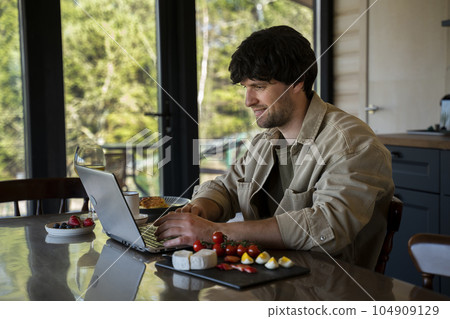 Smiling young man uses a laptop and has breakfast in the kitchen Smiling young man uses a laptop and has breakfast in the kitchen 104909129