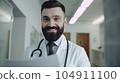 Hospital. Health care. Portrait of young smiling 30-year-old bearded caucasian man doctor wearing white medical coat with stethoscope around his neck, looking at camera. Hospital Corridor, AI 104911100