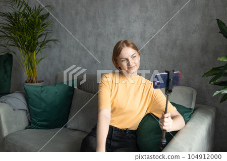 Cheerful young happy female blogger in casual comfortable outfit with braids waving and looking at screen while recording podcast on smartphone and sitting on sofa Cheerful young happy female blogger in casual comfortable outfit with braids waving and looking at screen while recording podcast on smartphone and sitting on sofa 104912000