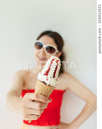 Happy cheerful girl in a red top and white sunglasses eats a colorful ice cream cone in her hands and smiles. 104912651