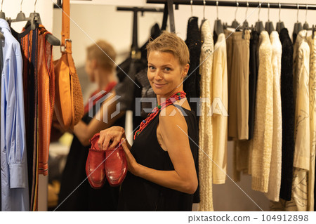 Adult stylish woman looking over the clothes at the boutique - holding red leather shoes Adult stylish woman looking over the clothes at the boutique - holding red leather shoes 104912898