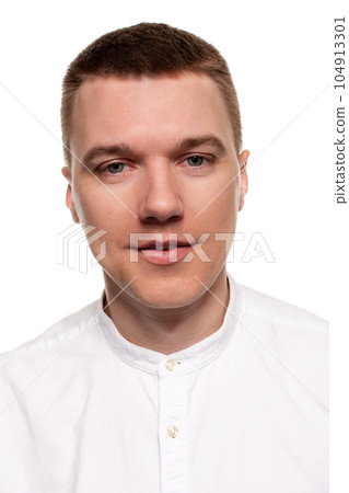 Charming handsome young man in a white shirt is making faces, while standing isolated on a white background Charming handsome young man in a white shirt is making faces, while standing isolated on a white background 104913301