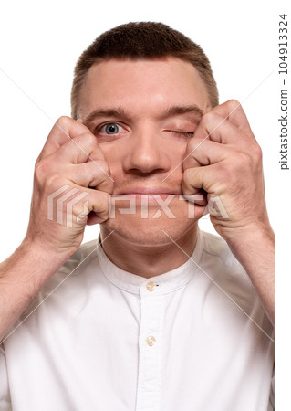 Charming handsome young man in a white shirt is making faces, while standing isolated on a white background 104913324