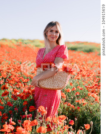 Attractive woman at a flowered poppy field. Beauty woman in red dress and straw handbag with wild flowers 104915659