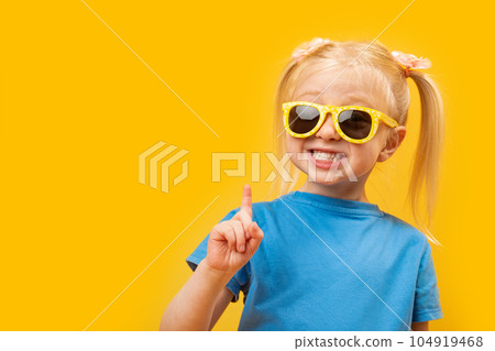 Smiling little blonde girl with two ponytails wears sunglasses and blue T-shirt with shows her index finger up. Copy space. 104919468