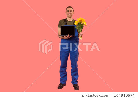 Full-length portrait of man in workers uniform holding laptop and bouquet of flowers, pink background. Copy space, mock up Full-length portrait of man in workers uniform holding laptop and bouquet of flowers, pink background. Copy space, mock up 104919674