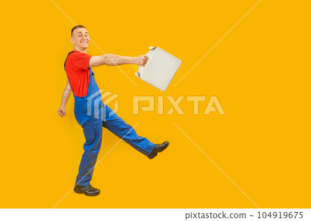 Man in blue jumpsuit and red T-shirt carries white cardboard box. Full-length portrait of courier on yellow background. Copy space, mock up. Man in blue jumpsuit and red T-shirt carries white cardboard box. Full-length portrait of courier on yellow background. Copy space, mock up. 104919675