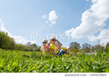 Little brother and sister wear sunglasses and sit on grass on blue sky background. Siblings bask in the sun. Country side. 104919805