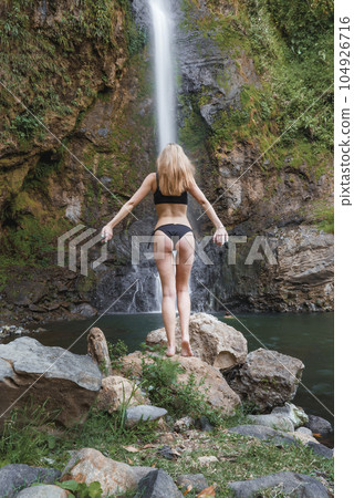Beautiful woman in bikini standing on rock admiring waterfall gushing through rocky hill in the rainforest jungle of Costa Rica 104926716