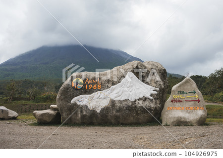 Rocks with sign of arenal 1968 trail and directional arrows in front of mountain volcano covered in clouds at Costa Rica 104926975