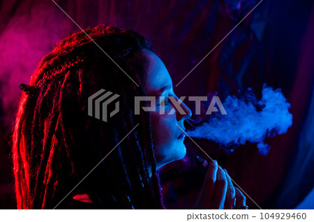 Profile of caucasian girl with dreadlocks smokes a vape in red blue light.  104929460
