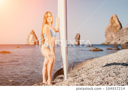 Woman sea sup. Close up portrait of happy young caucasian woman with long hair looking at camera and smiling. Cute woman portrait in a blue bikini posing on sup board in the sea 104937214