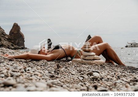 Woman bilds stones pyramid on seashore on a sunny day on the blue sea background. Happy holidays. Pebble beach, calm sea, travel destination. Concept of happy vacation on the sea, meditation, spa. Woman bilds stones pyramid on seashore on a sunny day on the blue sea background. Happy holidays. Pebble beach, calm sea, travel destination. Concept of happy vacation on the sea, meditation, spa. 104937320