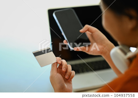 View over shoulder young woman holding credit card and using smart phone, shopping on Internet or payment online 104937558