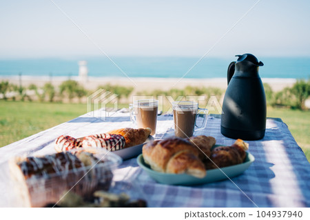 A picnic at the seaside beach. Breakfast with coffee, croissants and donuts by the ocean shore. 104937940