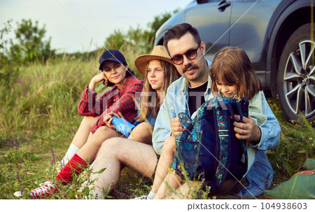 Loving family, man, woman and children sitting by the car on fitness matt and resting after active hiking on hills. 104938603