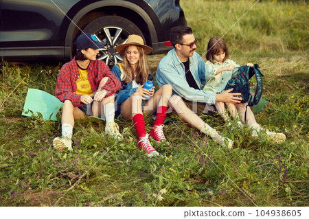 Family, man, woman and children sitting by the car on fitness matt and resting after active hiking on hills. Warm summer evening 104938605