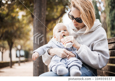 Mother sitting on bench in urban park, holding her infant baby boy child in her lap and feeding him baby food on nice spring sunny day. 104939337