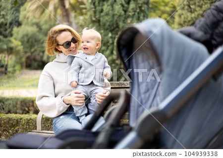 Mother sitting on bench in urban park, laughing cheerfully, holding her smiling infant baby boy child in her lap having baby stroller parked by their site. Mother sitting on bench in urban park, laughing cheerfully, holding her smiling infant baby boy child in her lap having baby stroller parked by their site. 104939338