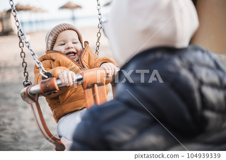 Mother pushing her cheerful infant baby boy child on a swing on sandy beach playground outdoors on nice sunny cold winter day in Malaga, Spain Mother pushing her cheerful infant baby boy child on a swing on sandy beach playground outdoors on nice sunny cold winter day in Malaga, Spain 104939339