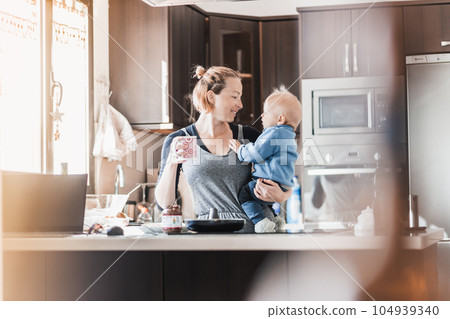 Happy mother and little infant baby boy together making pancakes for breakfast in domestic kitchen. Family, lifestyle, domestic life, food, healthy eating and people concept 104939340