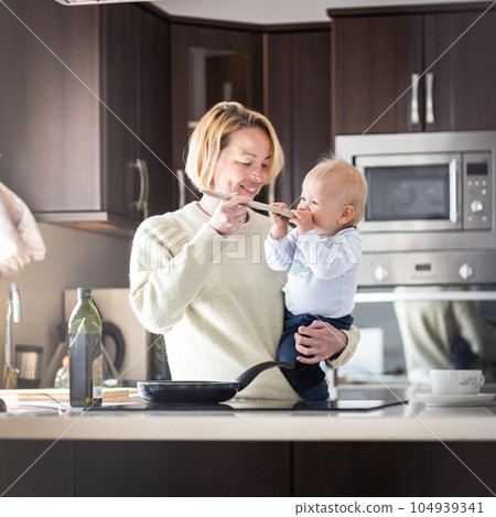 Happy mother and little infant baby boy cooking and tasting healthy dinner in domestic kitchen. Family, lifestyle, domestic life, food, healthy eating and people concept 104939341