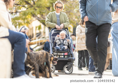 Mother walking and pushing his infant baby boy child in stroller in crowd of people wisiting sunday flea market in Malaga, Spain 104939342