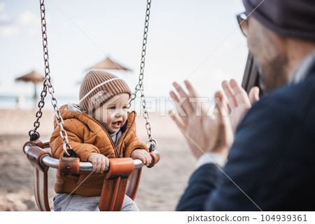 Father pushing hir cheerful infant baby boy child on a swing on sandy beach playground outdoors on nice sunny cold winter day in Malaga, Spain Father pushing hir cheerful infant baby boy child on a swing on sandy beach playground outdoors on nice sunny cold winter day in Malaga, Spain 104939361