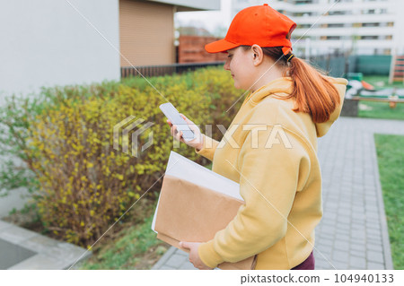 Happy Delivery woman with smartphone and parcel in hands front of the house. Courier send a package to destination. Happy professional shipping courier. Delivery postal service. Happy Delivery woman with smartphone and parcel in hands front of the house. Courier send a package to destination. Happy professional shipping courier. Delivery postal service. 104940133