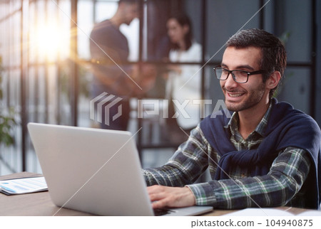 young businessman in glasses works with a computer remotely, sitting at the table. 104940875