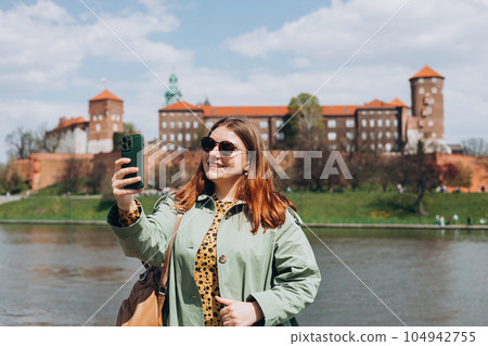 Attractive young female tourist is exploring new city. Redhead girl holding a smartphone in Krakow. Traveling. Happy girl walking in city and makes selfie poses against river and historic building Attractive young female tourist is exploring new city. Redhead girl holding a smartphone in Krakow. Traveling. Happy girl walking in city and makes selfie poses against river and historic building 104942755