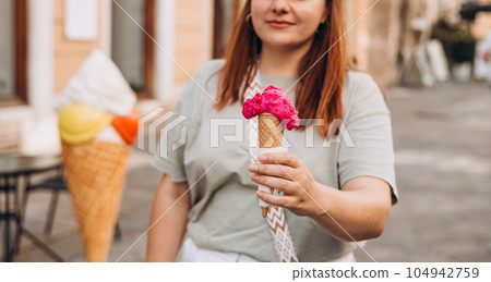 Colorful tasty ice cream cone in hand. Close up image of woman hand holding fresh waffle cone with pink ice cream Outdoors, food and rest concept 104942759