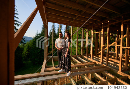 Man and woman inspecting their future wooden frame house nestled in the mountains near forest. Youthful couple at construction site in early morning. Concept of contemporary ecological construction. Man and woman inspecting their future wooden frame house nestled in the mountains near forest. Youthful couple at construction site in early morning. Concept of contemporary ecological construction. 104942880