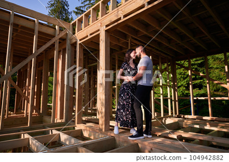 Man and woman inspecting their future wooden frame house nestled near the forest. Youthful couple at construction site in early morning. Concept of contemporary ecological construction. 104942882