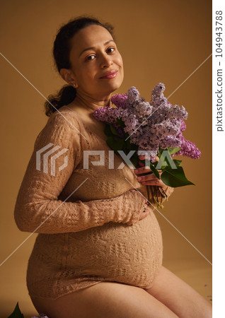 Gorgeous pregnant happy woman in fine beige knitted sweater, with blooming fragrant lilacs, smiles sweetly at the camera 104943788