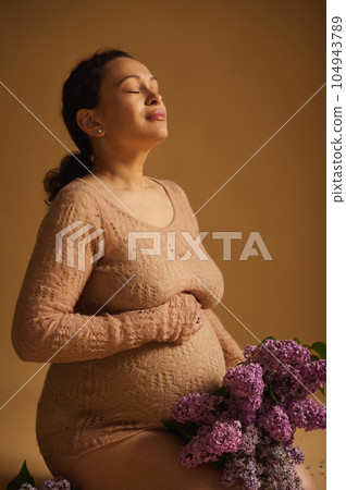 Gorgeous delightful gravid woman with fragrant lilacs, expectant mother strokes her belly, sits over beige background Gorgeous delightful gravid woman with fragrant lilacs, expectant mother strokes her belly, sits over beige background 104943789
